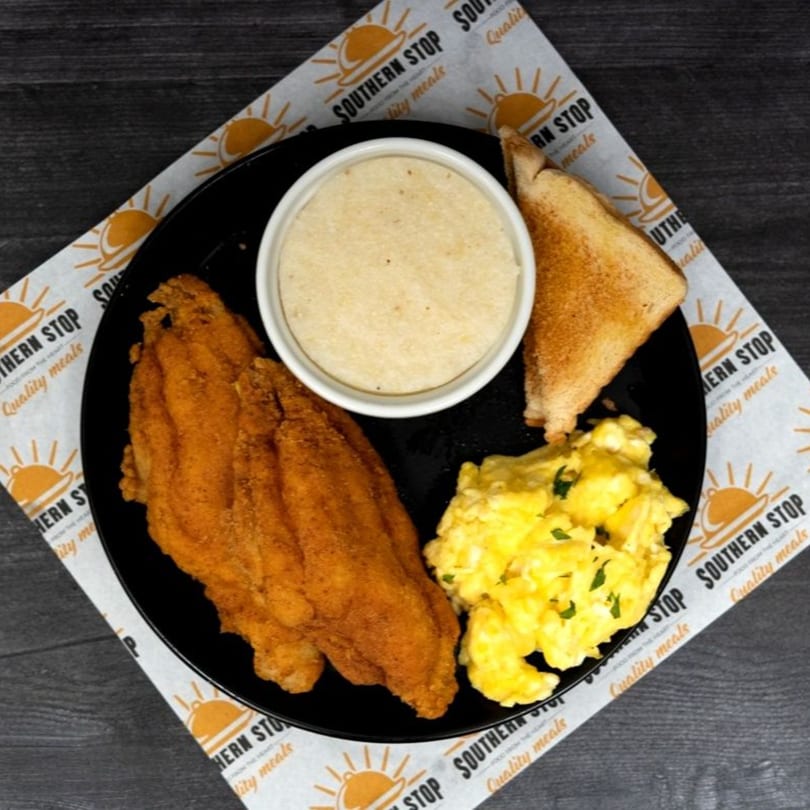 Southern Stop's fried fish, scrambled eggs, grits, and toast on a black plate.