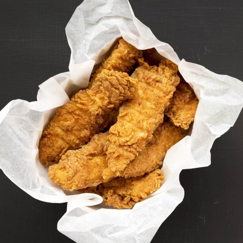 Crispy golden-brown fried chicken tenders, served in a white paper-lined basket on a dark surface.